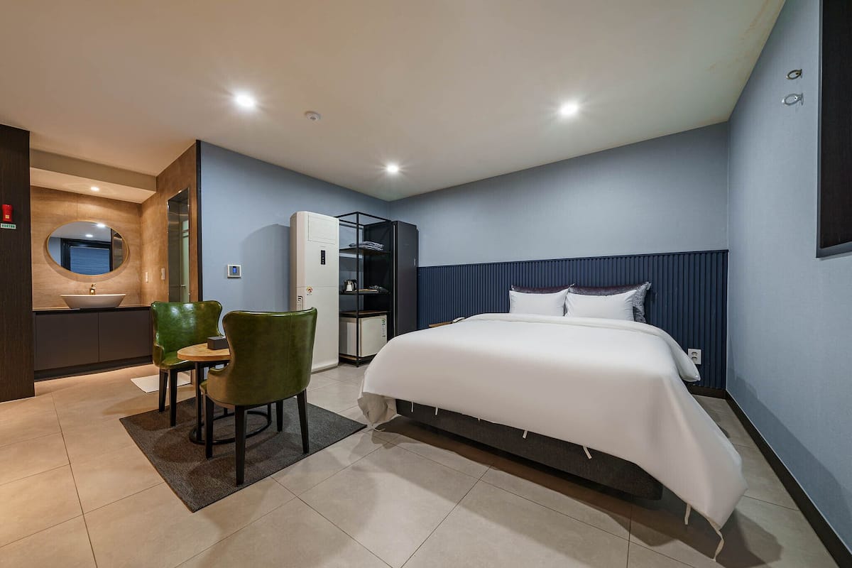A modern hotel room with a large bed, a small desk with a chair, a built-in wardrobe, and a bathroom with a sink and mirror.