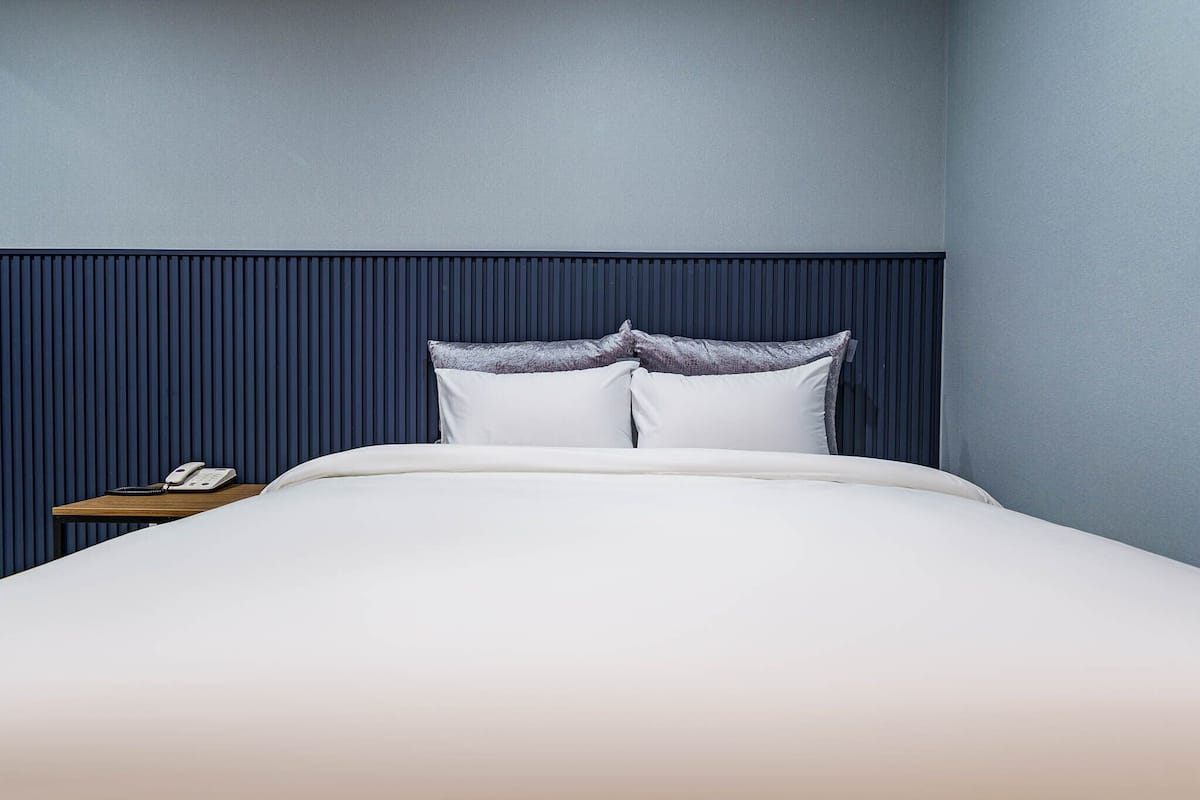 A neatly made bed with white linens and two pillows, a bedside table with a telephone, and a dark blue wall with vertical paneling.