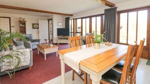 A spacious living room with a wooden dining table, comfortable seating, a television, and large windows. - Woodland View (Barnstaple)