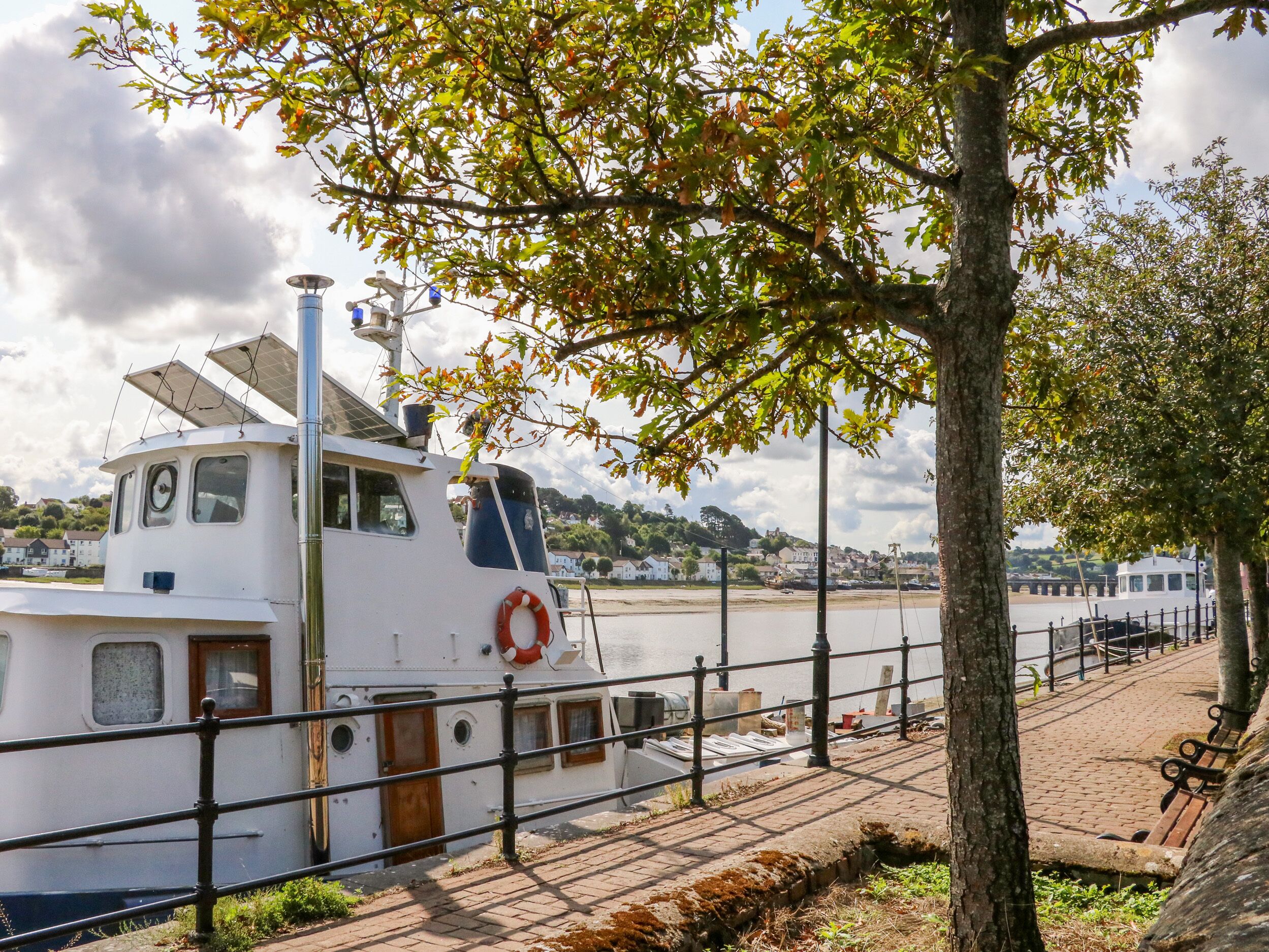 A white boat docked at a pier with a lifebuoy attached, surrounded by a brick walkway and a metal railing.