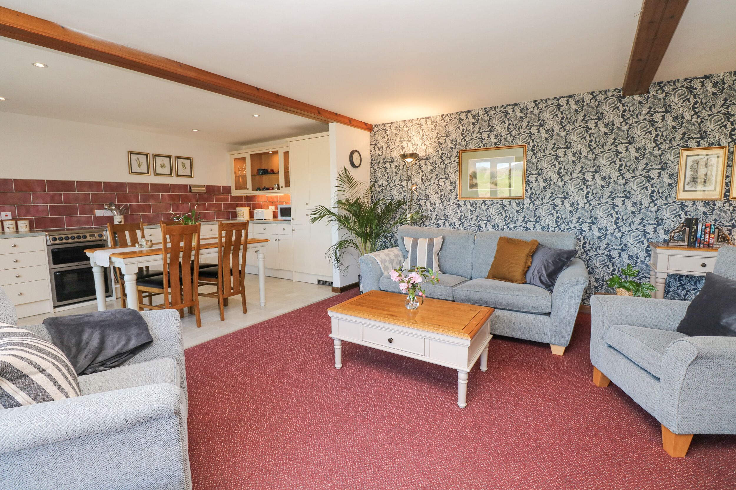 A living room with a red carpet, grey sofas, a wooden coffee table, and a kitchen in the background.