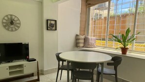 A living room with a round dining table, four chairs, a flat-screen TV on a white cabinet, a clock on the wall, and a window with a view of a fence and greenery.