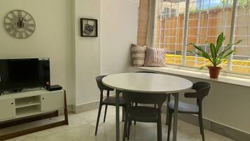 A living room with a round dining table, four chairs, a flat-screen TV on a white cabinet, a clock on the wall, and a window with a view of a fence and greenery.