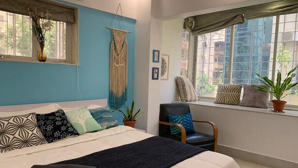 A bedroom with a bed, a chair, a macrame wall hanging, and a window with a view of buildings.