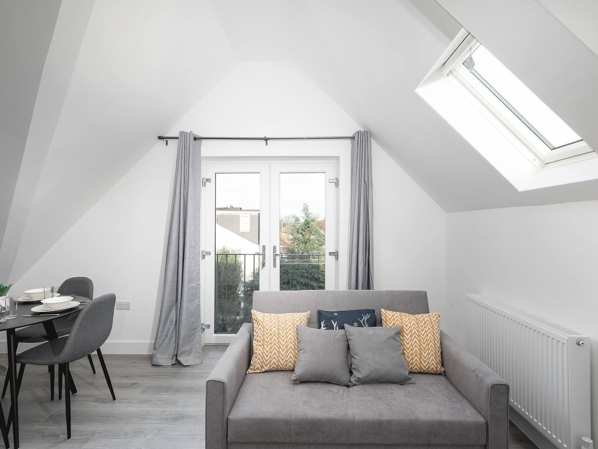 Une chambre de style loft moderne, dotée d’une lucarne, d’un canapé gris agrémenté de coussins à motifs, d’un coin repas avec une table et des chaises, et offrant une vue à travers une porte vitrée.