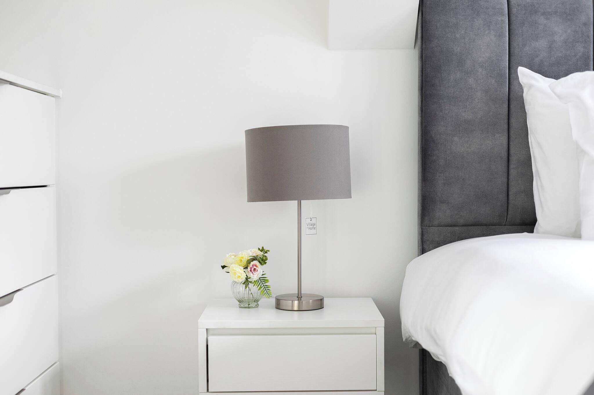 A white bedside table with a lamp, a vase with flowers, and a bed with white bedding.