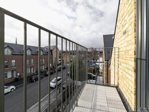 Un balcon avec des balustrades en métal donnant sur une rue où sont garées des voitures et où se trouvent des immeubles d’habitation.