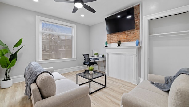 Un salon moderne doté d’un ventilateur de plafond, d’un téléviseur à écran plat fixé sur un mur en briques, d’une cheminée et d’une grande fenêtre avec des stores.