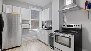 Une cuisine moderne avec des meubles blancs, un réfrigérateur en acier inoxydable, un four à micro-ondes, une cuisinière et une fenêtre avec des stores.