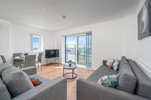 A modern living room with a grey sofa, a TV, a dining table, and a view of a cityscape through large windows. - Skyvillion-apartments Near River Thames Excel & O2 Woolwich (London)