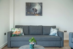 A grey sofa with two patterned pillows, a small round table with a plant, and a painting of an elephant on the wall. - Skyvillion-apartments Near River Thames Excel & O2 Woolwich (London)