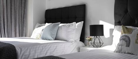 A hotel room with two beds, a large window with curtains, a wall-mounted artwork, a bedside table with a lamp, and decorative pillows.
