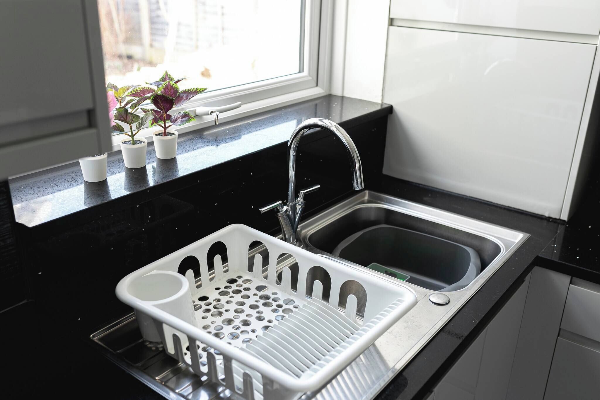 A kitchen with a black countertop, a stainless steel sink, a white dish rack, and a window with potted plants.