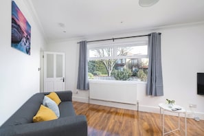 A modern living room with a grey sofa, a small white table with a vase, a large window with grey curtains, and a white radiator. - Skyvillion - Enfield 4bed House W Garden & Parking (Enfield)