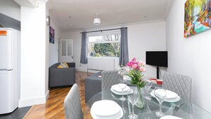A modern living room with a dining table set for two, a grey sofa, and a large window with curtains. - Skyvillion - Enfield 4bed House W Garden & Parking (Enfield)