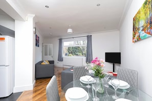 A modern living room with a dining table set for two, a grey sofa, and a large window with curtains. - Skyvillion - Enfield 4bed House W Garden & Parking (Enfield)