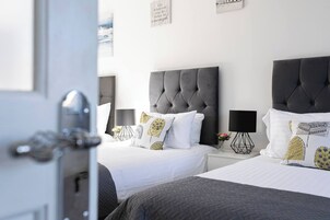 Habitación de hotel con dos camas, cada una con cabecero capitonado, ropa de cama blanca y cojines decorativos. Hay una mesita de noche con una lámpara y un florero con flores.