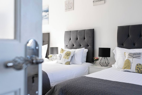Ett hotellrum med två sängar, var och en med ett stoppat sänggavel, vita sängkläder och dekorativa kuddar. Det finns ett nattduksbord med en lampa och en vas med blommor.