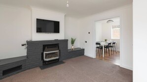A living room with a fireplace, a flat-screen TV mounted above it, and a dining area with a table and chairs. - Skyvillion - 3bed Home W Grdn For A Family Getaway (Stevenage)