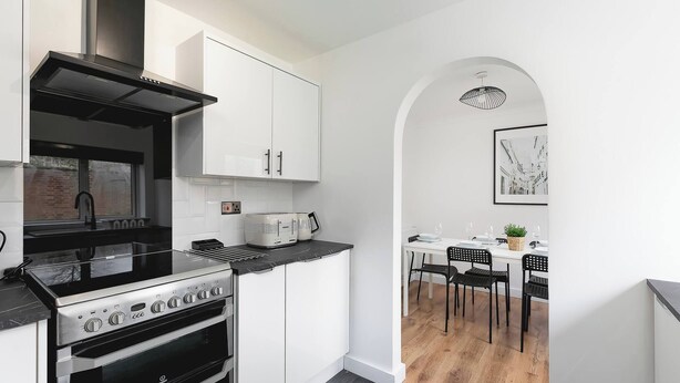 A modern kitchen with white cabinetry, a black stove, and a dining area with a white table and chairs. - Skyvillion - 3bed Home W Grdn For A Family Getaway (Stevenage)