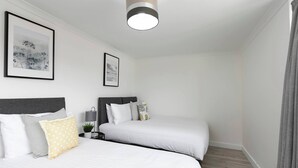 A hotel room with two beds, a ceiling light, framed artwork on the wall, and a nightstand with a lamp. - Skyvillion - 3bed Home W Grdn For A Family Getaway (Stevenage)