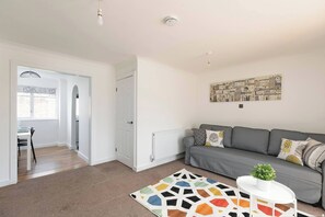 Ein modernes Wohnzimmer mit einem grauen Sofa, einem farbenfrohen Teppich und einem weißen Tisch mit einer Pflanze.