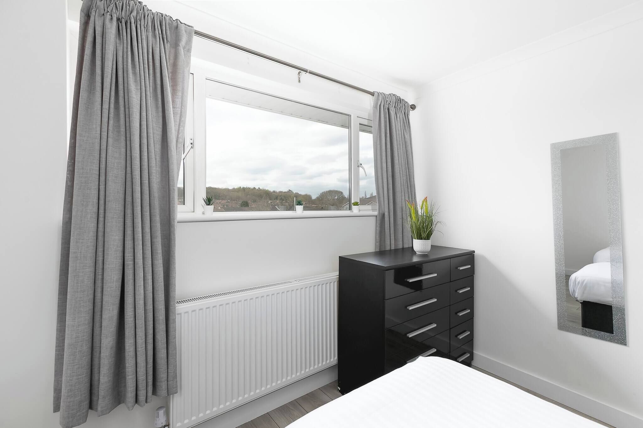 A bedroom with a window, a radiator, a black dresser, a potted plant, and a mirror on the wall.