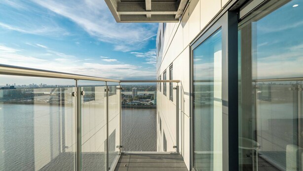 A modern building with large glass windows, a balcony with a metal railing, and a view of a body of water and distant cityscape.