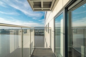 Ein modernes Gebäude mit großen Glasfenstern, einem Balkon mit Metallgeländer und Blick auf ein Gewässer sowie eine entfernte Stadtlandschaft.