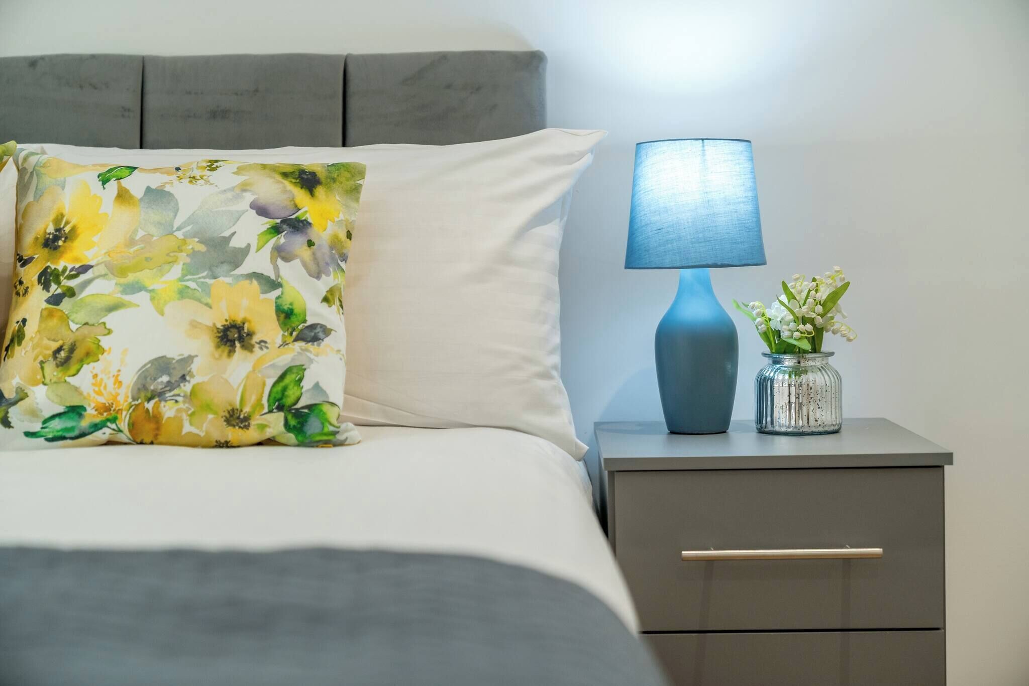 A neatly made bed with a floral pillow, a bedside lamp, and a small vase with flowers on a nightstand.