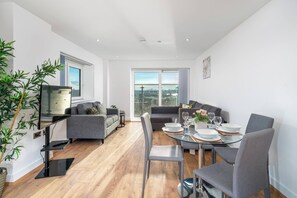 Ein modernes Wohnzimmer mit Essbereich, einem Flachbildfernseher, grauen Sofas und einem großen Fenster mit Blick auf die Stadt.