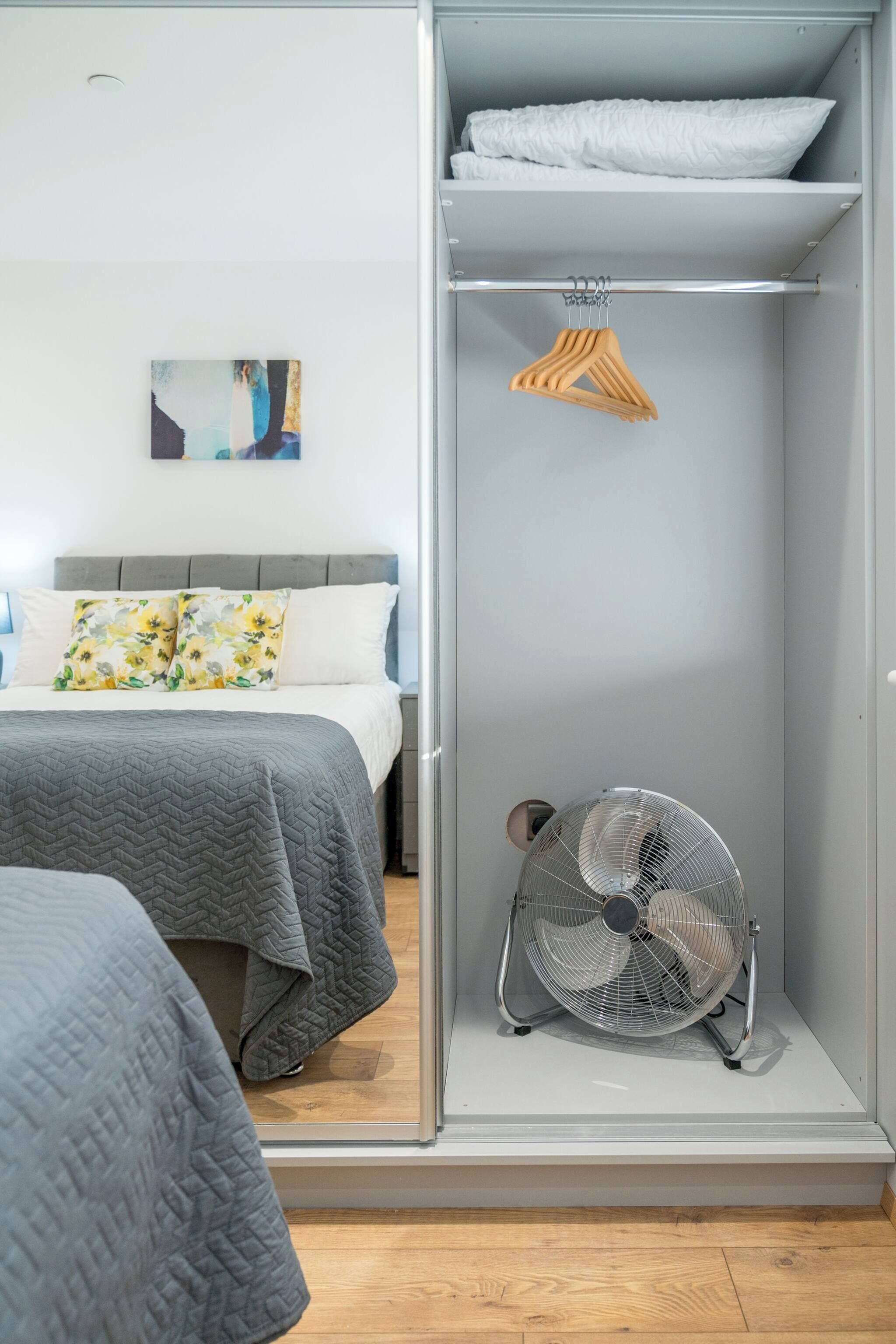 A bedroom with a bed, a grey quilted bedspread, a grey wall, a white pillow, a white pillow with a floral design, a grey fan, and a wooden hanger.