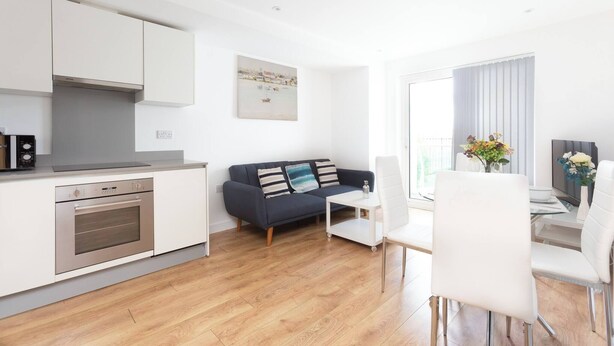 A modern kitchen with built-in appliances, a dark blue sofa with striped pillows, a dining area with white chairs, and a large window with curtains. - Skyvillion - Tower Point Enfield 1bed With Balcony (Enfield)
