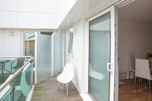 Un balcón con barandilla de vidrio, una silla blanca y una zona de comedor con sillas y una mesa.
