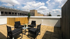 An outdoor patio with a table and chairs, a potted plant, and a view of a building with a wooden fence and a metal railing. - Skyvillion - Huge 4bed Apartment Wbalcony - Parking (Enfield)