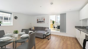 A modern apartment with a dining area, a gray sofa, a kitchen, and a large window. - Skyvillion - Huge 4bed Apartment Wbalcony - Parking (Enfield)
