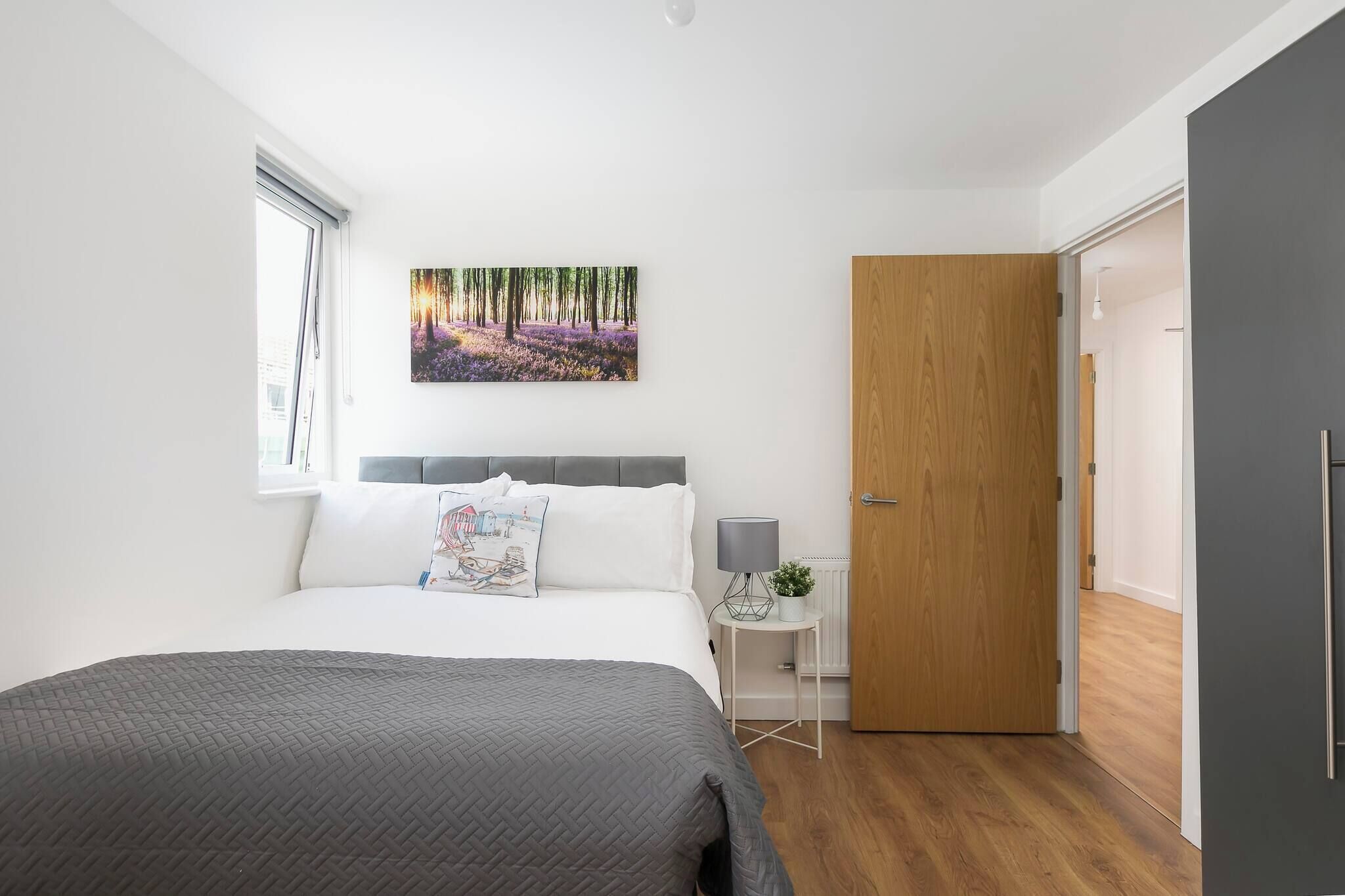Un dormitorio con cama, mesita de noche, lámpara, puerta y un cuadro colgado en la pared.