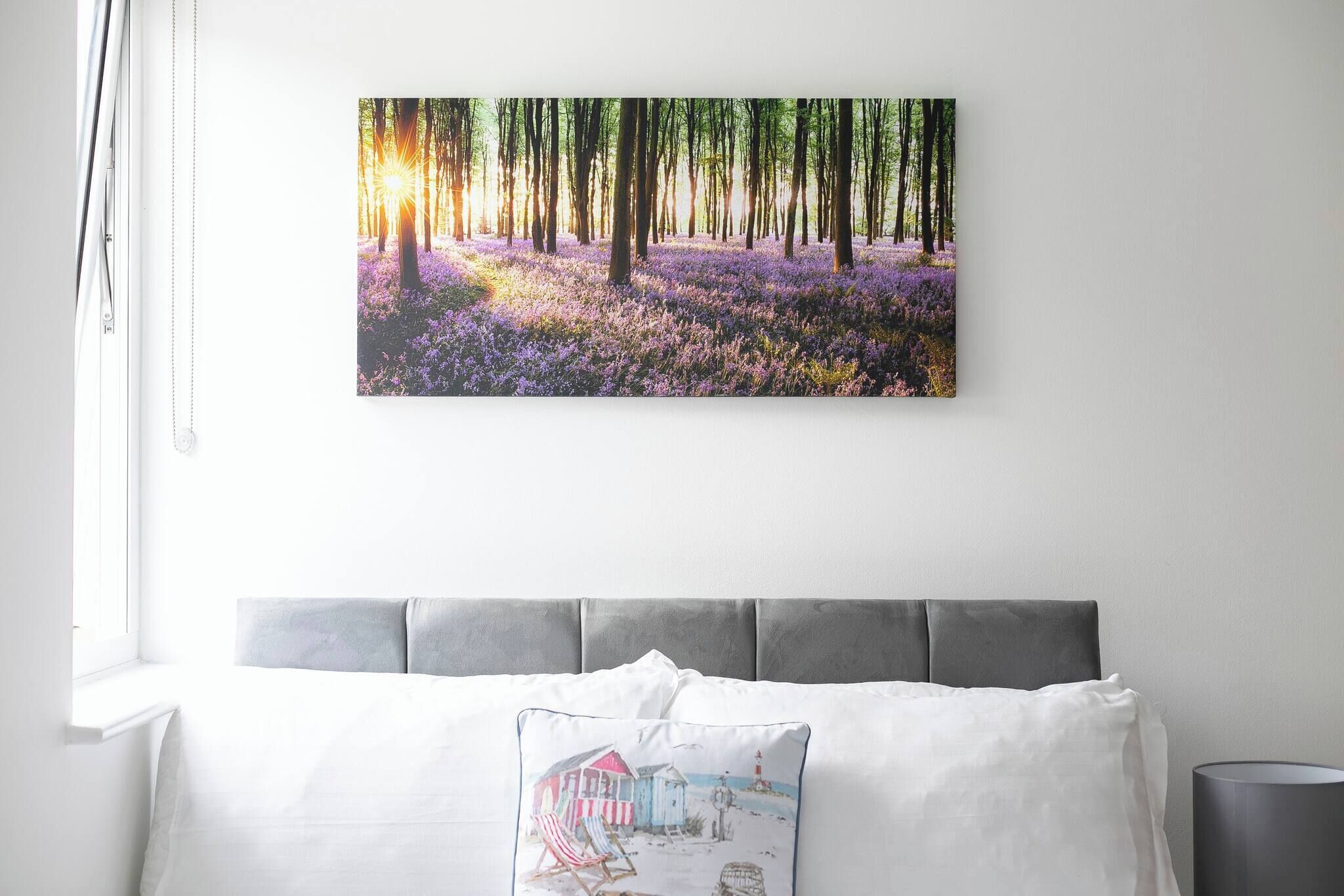 A bedroom with a bed, a pillow with a beach scene, and a wall-mounted painting of a forest.