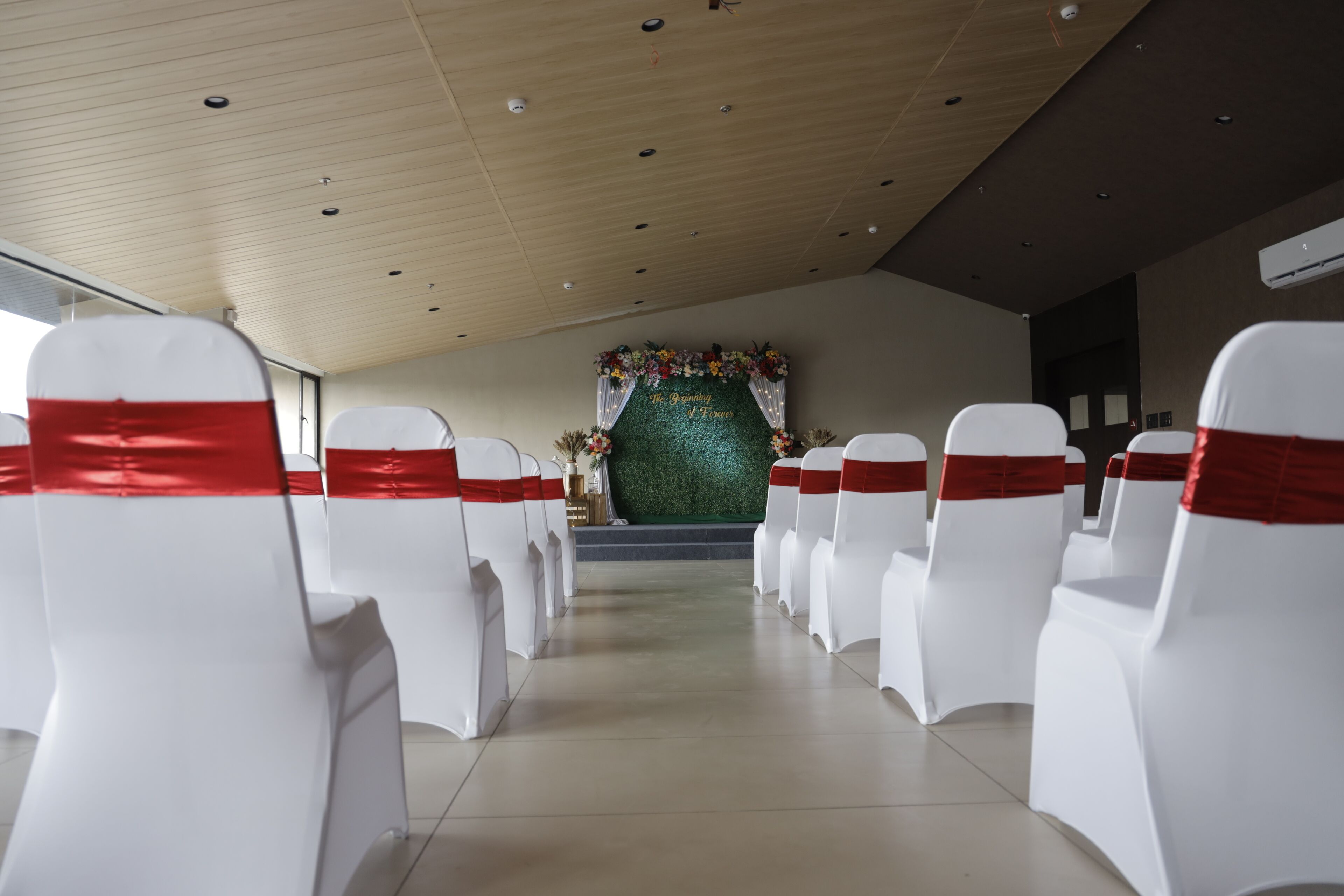 A church interior with rows of white chairs with red sashes, a floral altar backdrop, and a wooden ceiling.