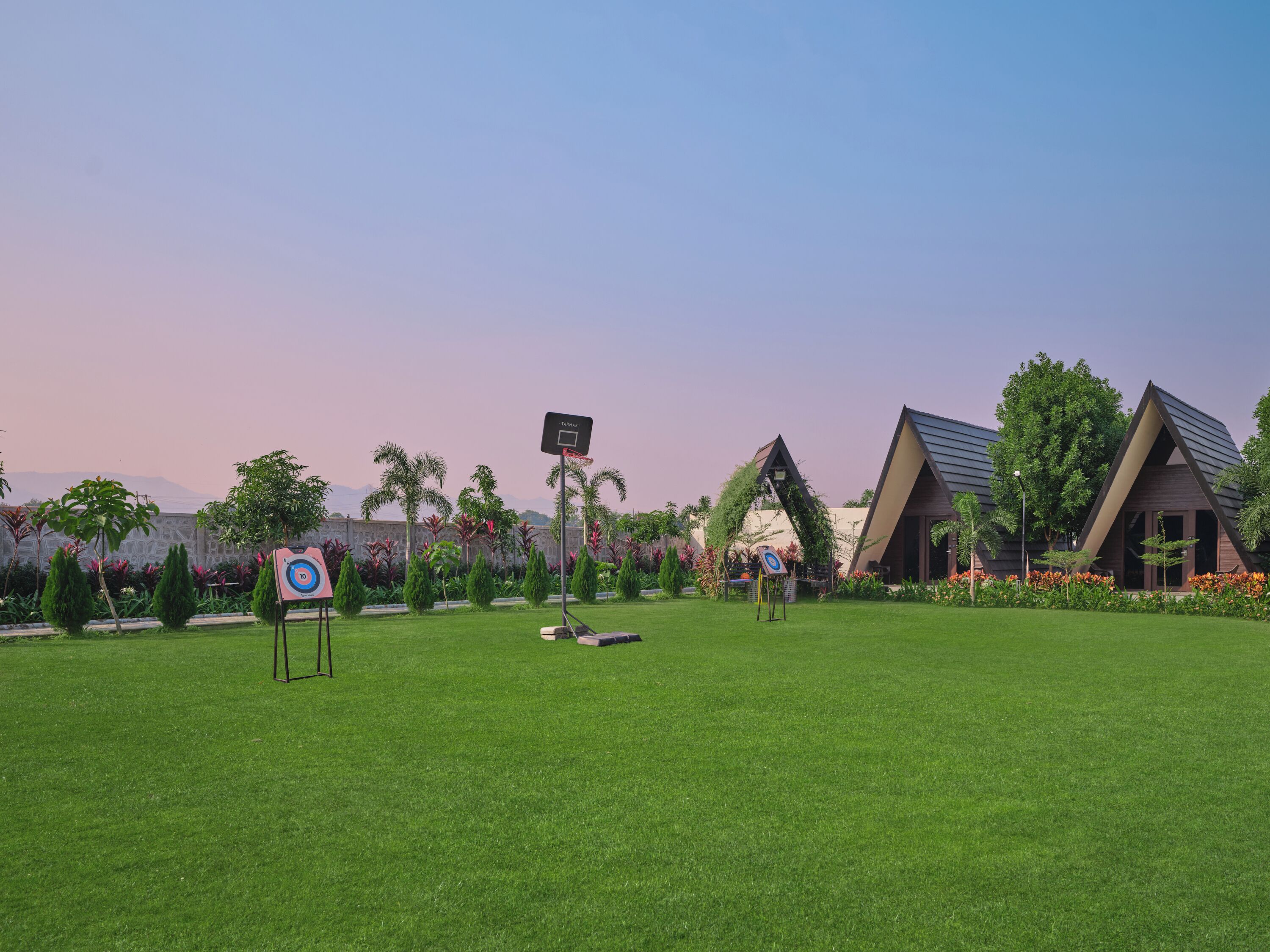 A well-maintained lawn with a basketball hoop and a target sign, surrounded by lush greenery and modern buildings with triangular roofs.