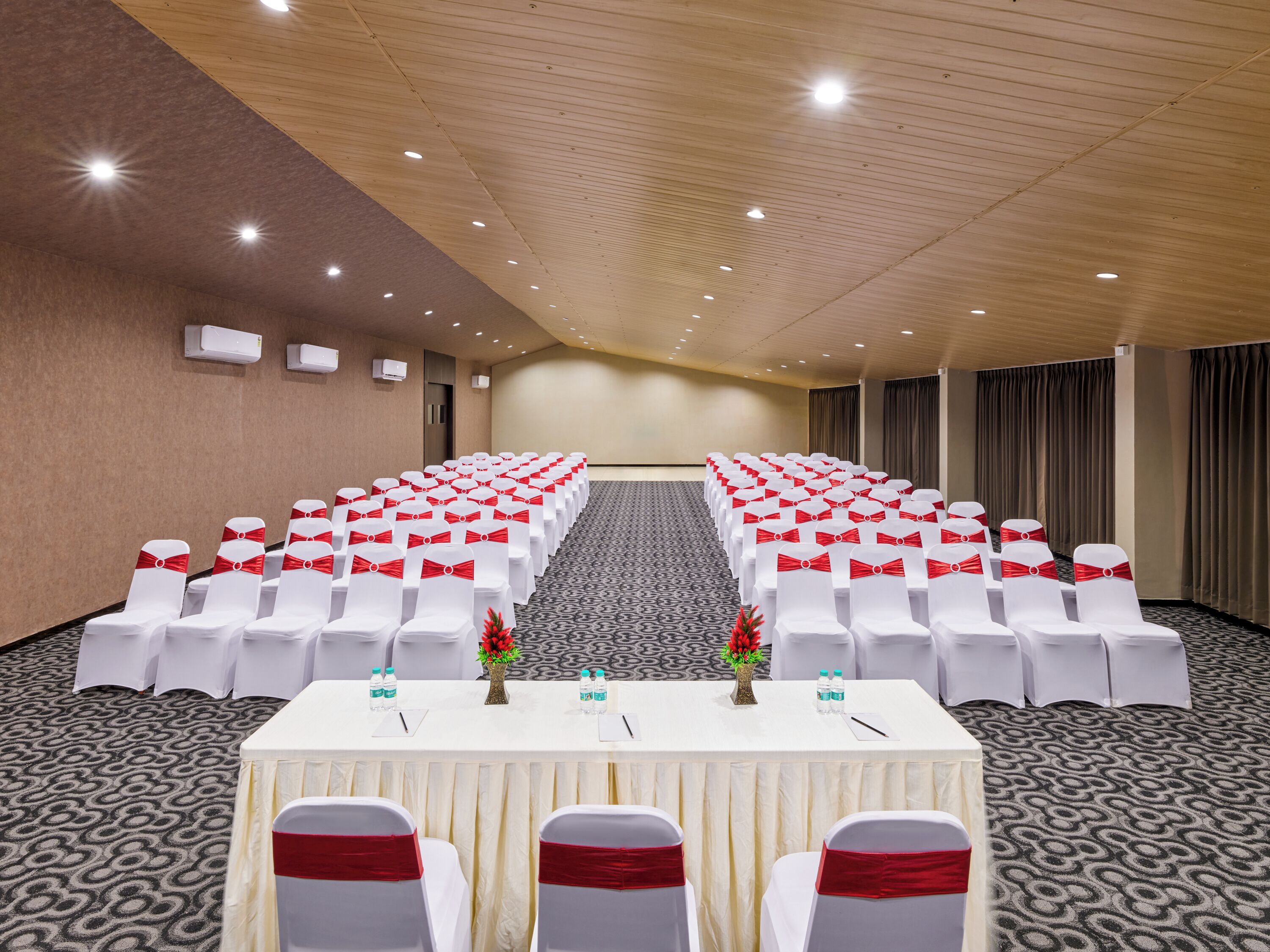 Un salón de conferencias con filas de sillas blancas, cintas rojas y mesas con botellas de agua.