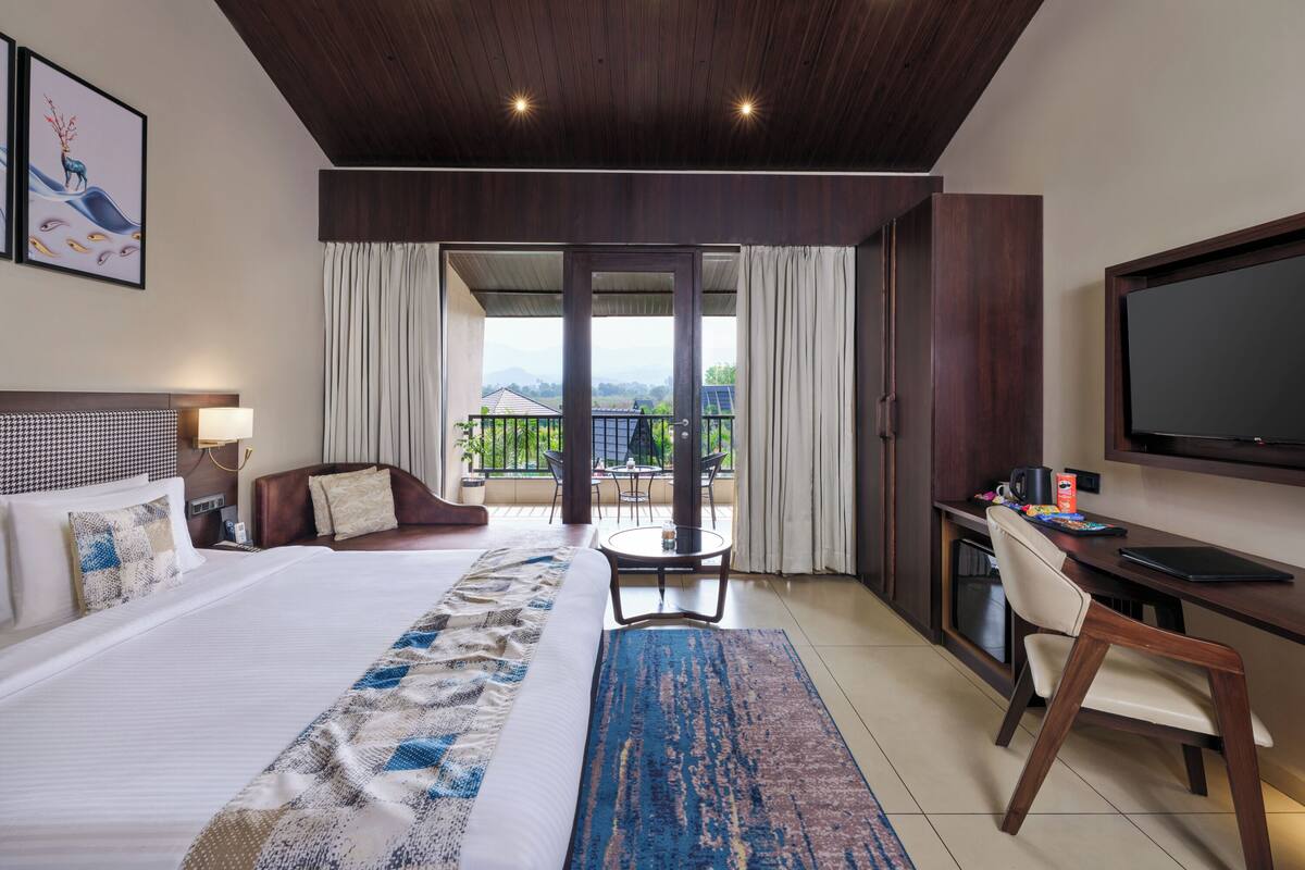 A hotel room with a large bed, a desk, a chair, a TV, and a balcony with a view.