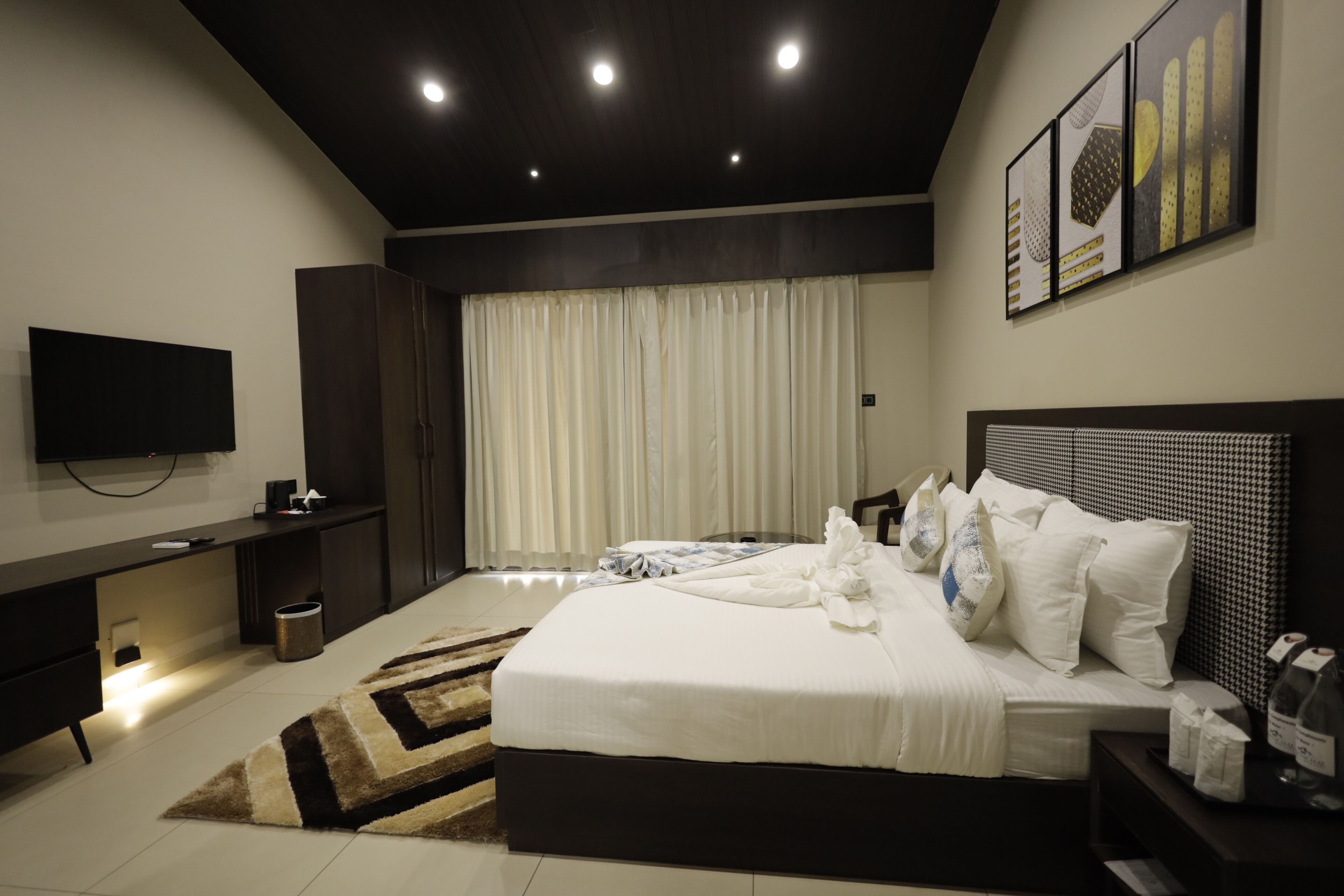 Un dormitorio moderno con una cama grande, un televisor de pantalla plana montado en la pared, un escritorio y un amplio ventanal con cortinas.