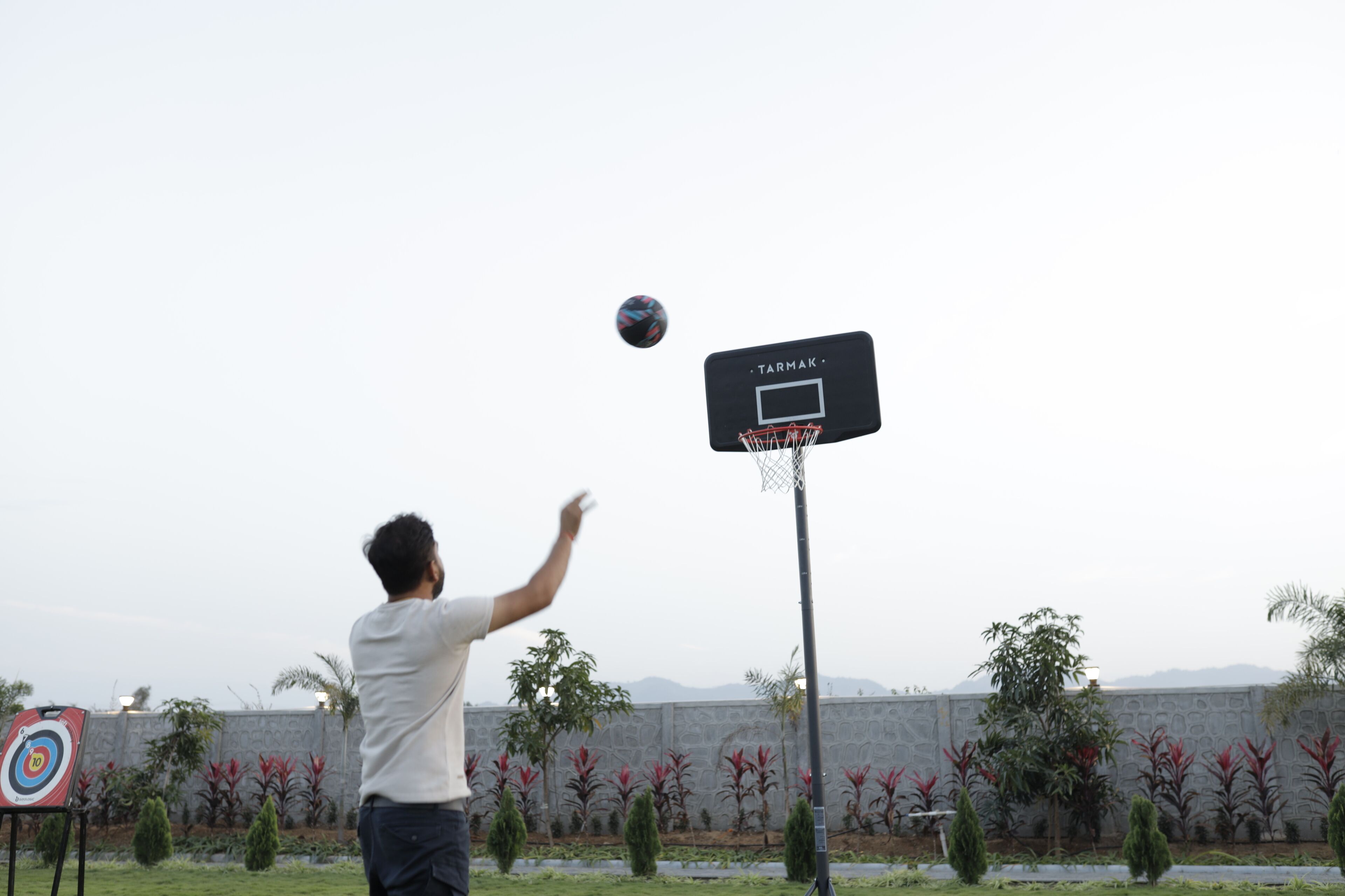 A basketball hoop with a target on the ground, a man in a white shirt and dark pants, and a landscaped area with trees and a low wall in the background.