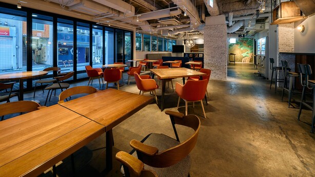 A modern restaurant interior with high ceilings, exposed ductwork, and large windows offering views of the street. Tables and chairs are arranged for dining. - Hotel Plus Hostel TOKYO KAWASAKI (Kawasaki)