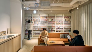 A modern interior with a kitchen area, a seating area with a sofa and a coffee table, and a large bookshelf filled with books. - Hotel Plus Hostel TOKYO KAWASAKI (Kawasaki)