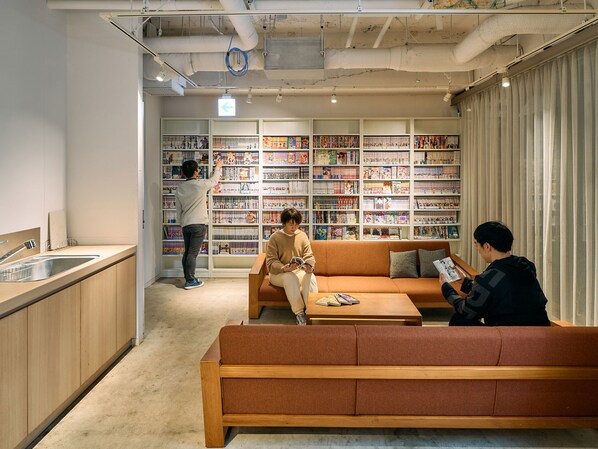 A modern interior with a kitchen area, a seating area with a sofa and a coffee table, and a large bookshelf filled with books. - Hotel Plus Hostel TOKYO KAWASAKI (Kawasaki)