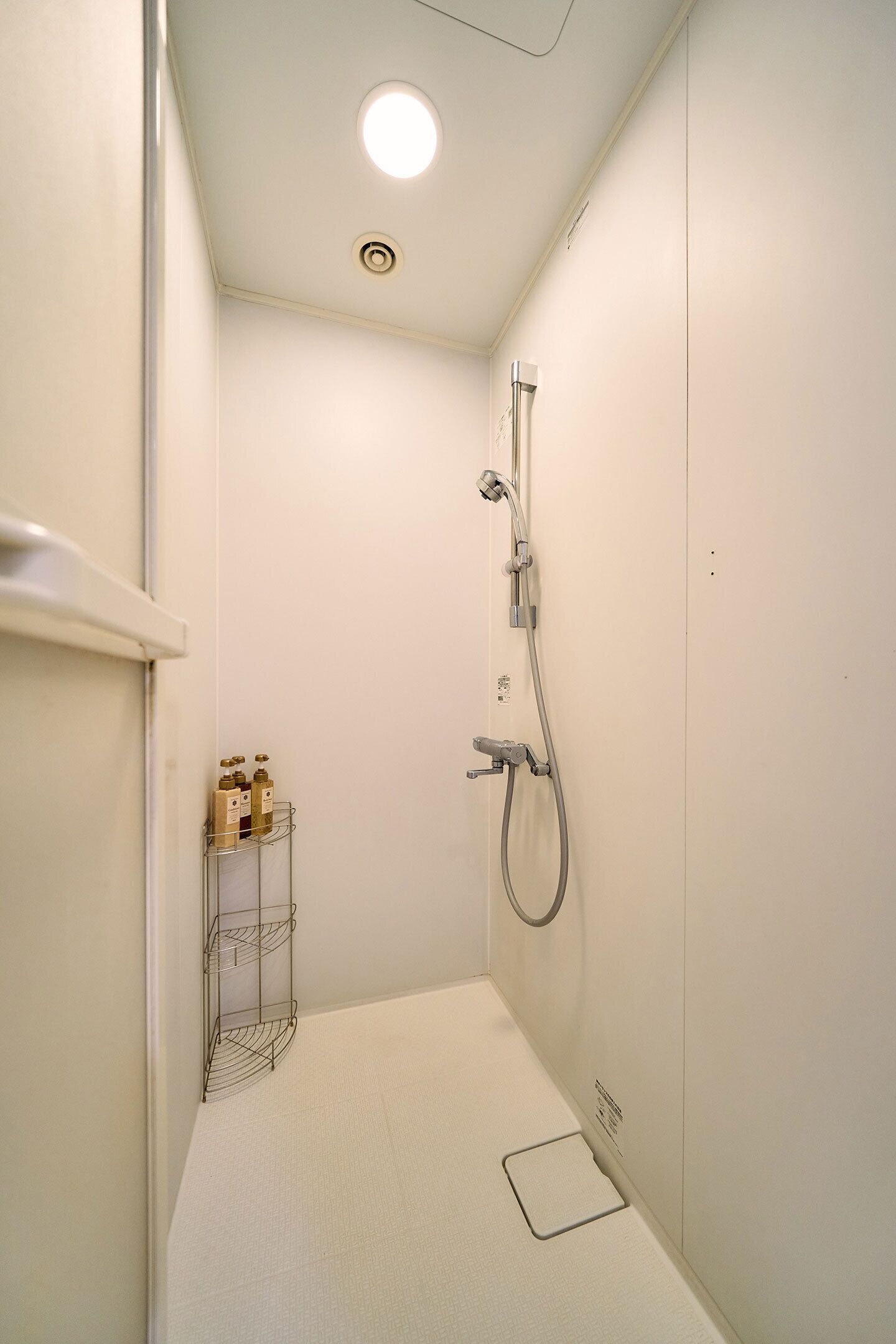 Un baño pequeño y blanco con ducha, un perchero con botellas y una lámpara en el techo.