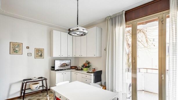 A dining area with a white table and chairs, a small kitchen area with cabinets, a television, and a window with curtains.
