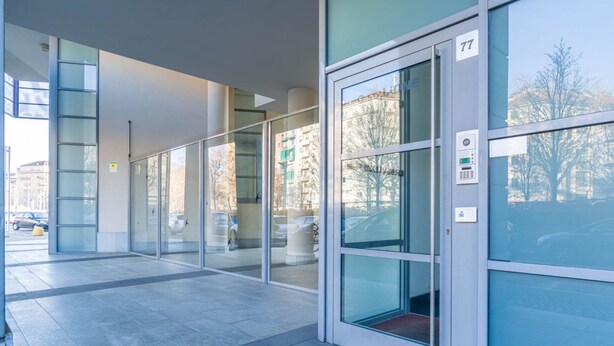 Entrada de un edificio moderno con puerta de vidrio, un letrero con número y un teclado digital.